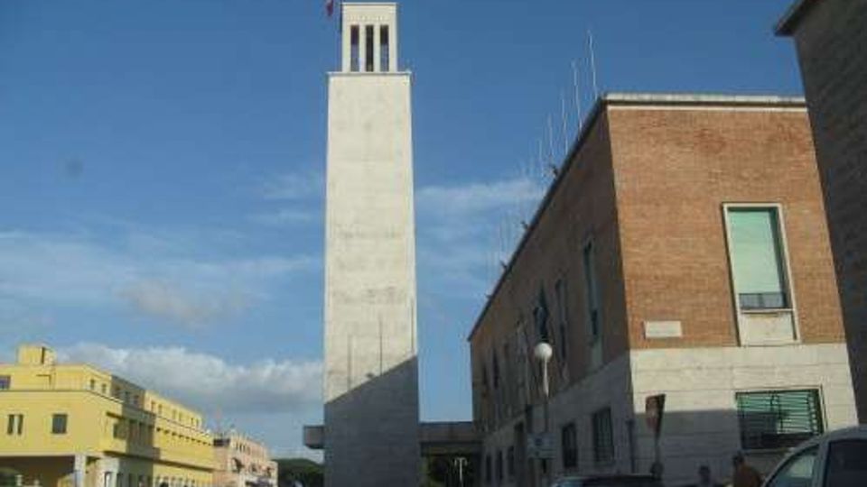 Strada urbana con edifici in mattoni e intonaco e una torre alta e rettangolare in cemento con bandiera in cima. Auto parcheggiate e in transito ai lati della via, con cielo azzurro sullo sfondo.