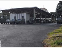 Ampia tettoia industriale in metallo con struttura a travi e pilastri, affiancata da un edificio prefabbricato a pannelli. Sotto la copertura sono parcheggiati diversi veicoli e mezzi, con grande piazzale asfaltato e alberi sullo sfondo.