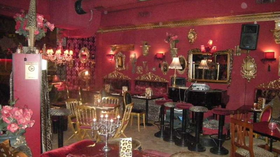 Interno di un locale notturno o lounge con pareti rosso scuro, specchi e decorazioni dorate, luci soffuse e lampadari. Tavolini e sedute imbottite con motivi leopardati, divanetti lungo le pareti e un’area con sgabelli e impianto audio vicino a una parete con specchi.