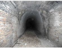 Interno di un tunnel sotterraneo in mattoni con volta a botte, pareti grezze e pavimento coperto di ghiaia e detriti. Il corridoio si restringe in prospettiva fino a un’area buia in fondo.