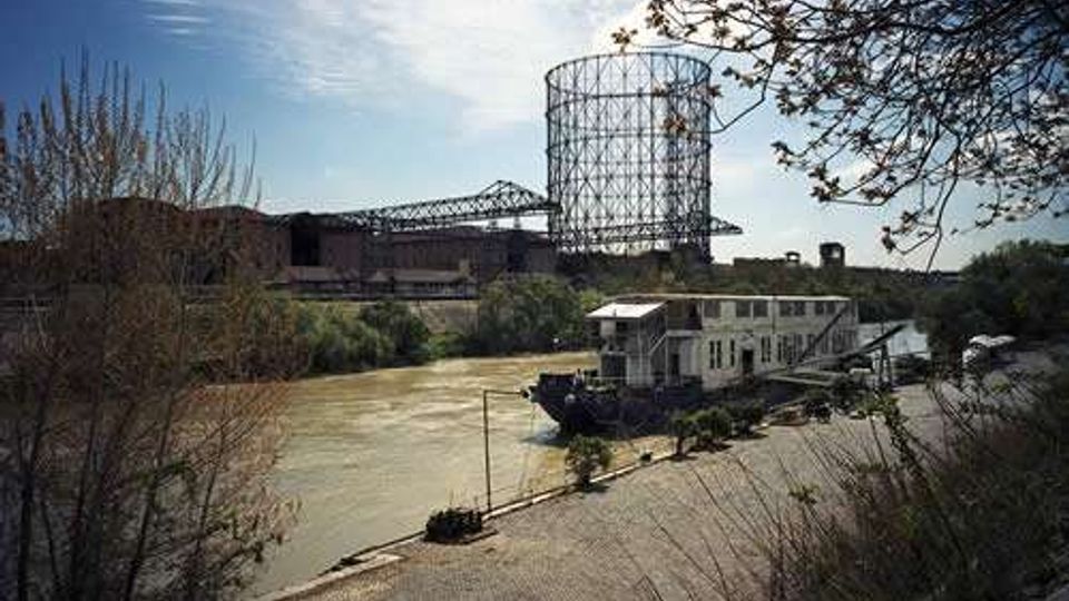 Vista di un fiume con un percorso pedonale sulla riva e vegetazione ai margini; in primo piano si vede una struttura galleggiante ormeggiata. Sullo sfondo compaiono edifici industriali e una grande torre cilindrica a traliccio in metallo.
