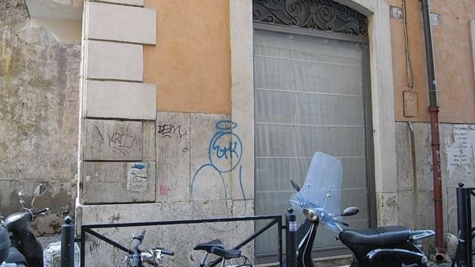 Facciata di un edificio urbano con intonaco ocra e basamento in pietra, caratterizzata da una grande porta ad arco con cornice chiara e grata decorativa. Davanti, su strada in sampietrini, sono parcheggiati scooter e biciclette vicino a una ringhiera metallica e a paletti.