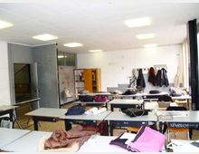 Aula scolastica luminosa con file di banchi e sedie, soffitto a pannelli con luci al neon e pavimento in graniglia. Sul fondo si vedono pareti bianche con appendiabiti e giacche appese, scaffali e una porta aperta verso un corridoio.