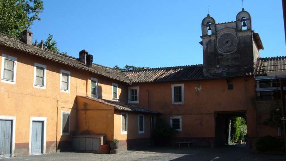 Cortile lastricato circondato da edifici intonacati color ocra con finestre e tetti in coppi, con un grande portale ad arco che conduce all’esterno. Sullo sfondo si vede una struttura tipo campanile con due piccole campane e un orologio sotto un cielo sereno.