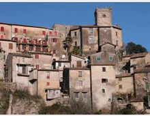 Borgo medievale su un pendio con case in pietra addossate tra loro, finestre e persiane colorate e tetti in coppi. In alto domina una torre quadrata merlata in pietra, con cielo sereno sullo sfondo.