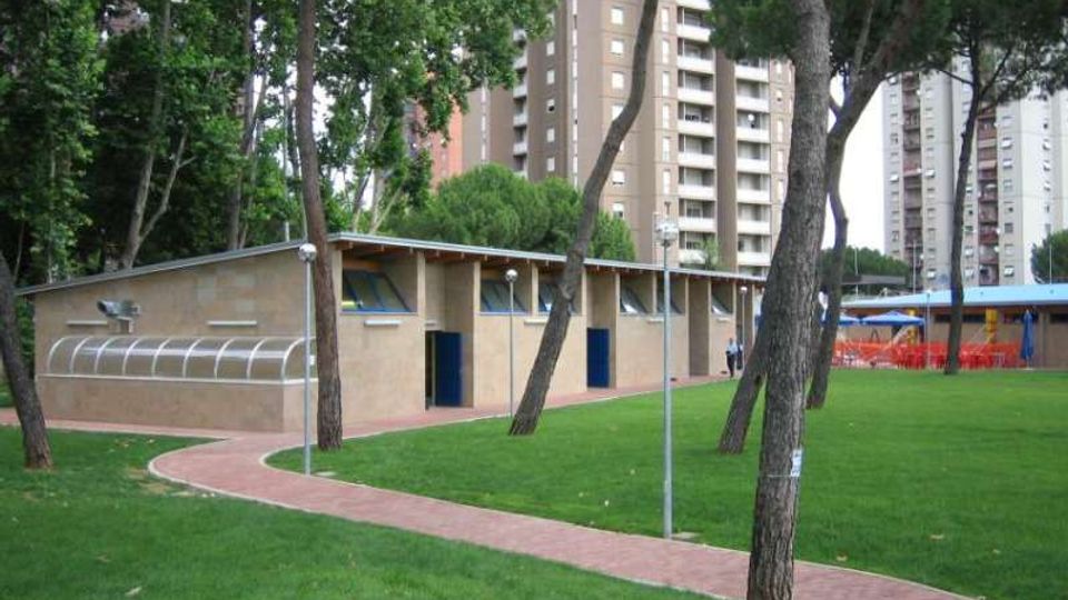 Edificio basso in muratura con tetto inclinato e porte blu, situato in un parco con prato verde, alberi alti e un sentiero pavimentato curvo in primo piano. Sullo sfondo si vedono alti palazzi residenziali e alcuni arredi esterni coperti.