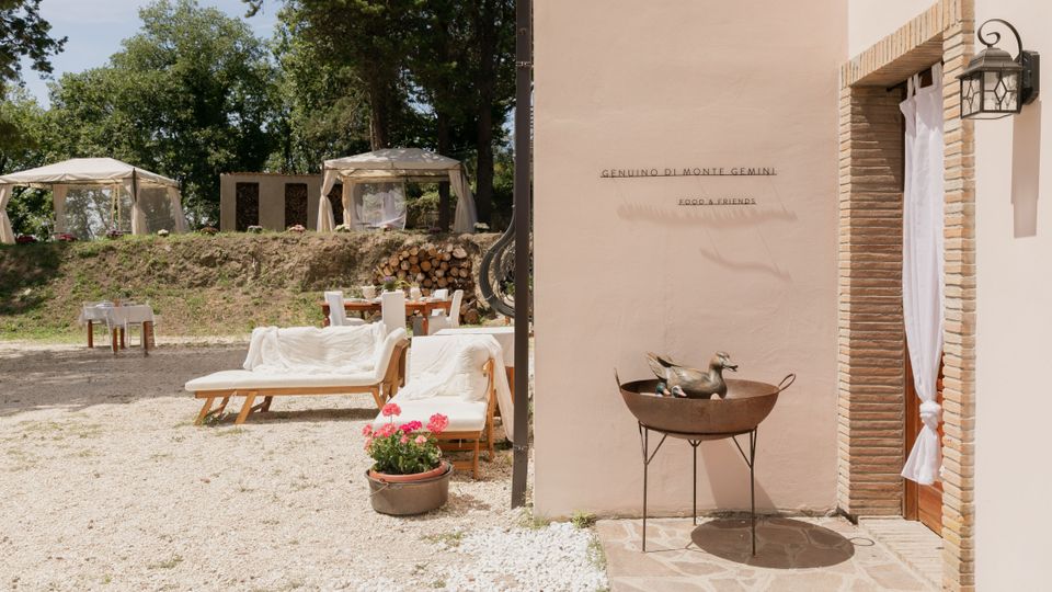 Cortile esterno di una struttura rustica con parete in intonaco chiaro e porta incorniciata in mattoni, illuminata da una lanterna. Davanti ci sono lettini e tavoli su ghiaia, una catasta di legna e gazebo bianchi tra alberi sullo sfondo.