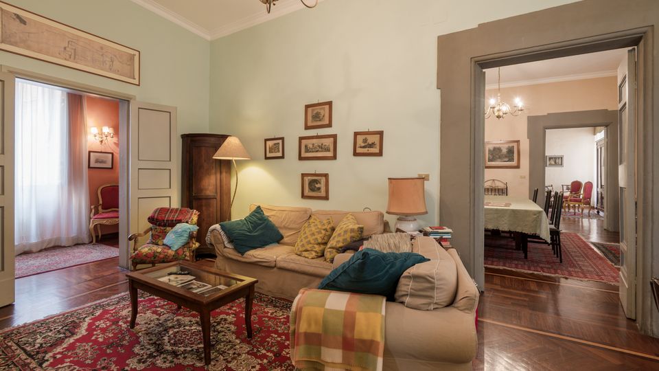 Soggiorno in stile classico con divani beige, tavolino basso e grande tappeto rosso su pavimento in parquet. Pareti verde chiaro con quadri incorniciati, lampadario a soffitto e ampie porte che collegano a stanze adiacenti con arredi coordinati.