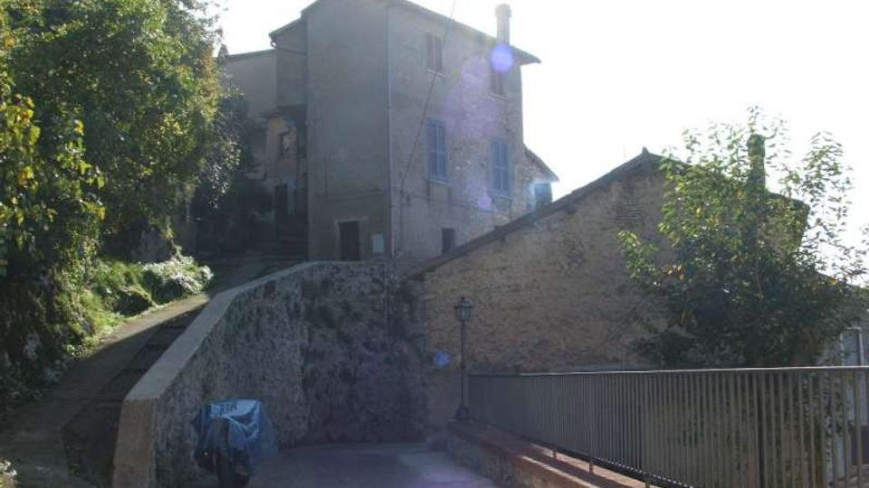 Strada in lieve salita con pavimentazione in pietra che conduce a edifici in muratura su un terrapieno, con un alto muro di contenimento. A destra c’è una ringhiera metallica lungo il bordo e vegetazione con alberi sui lati.