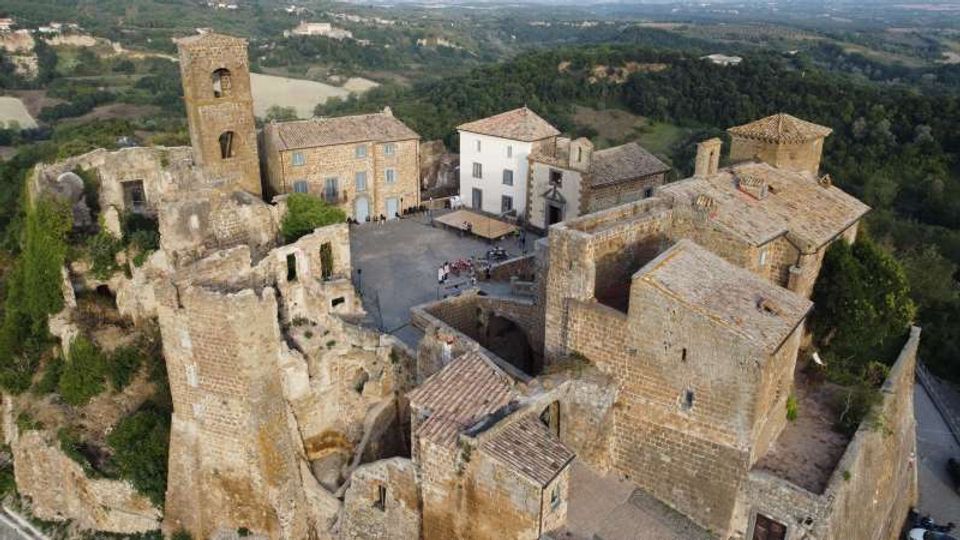 Veduta aerea di un borgo medievale in pietra con rovine di mura e torri, una piazza centrale e edifici restaurati con tetti in coppi. Intorno si estendono boschi e campi collinari, con una strada che costeggia il complesso sul lato destro.