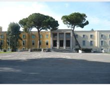 Edificio pubblico a due piani con facciata simmetrica e ingresso centrale incorniciato da colonne e una breve scalinata. Davanti si apre un ampio piazzale asfaltato con due grandi pini marittimi ai lati dell’entrata.