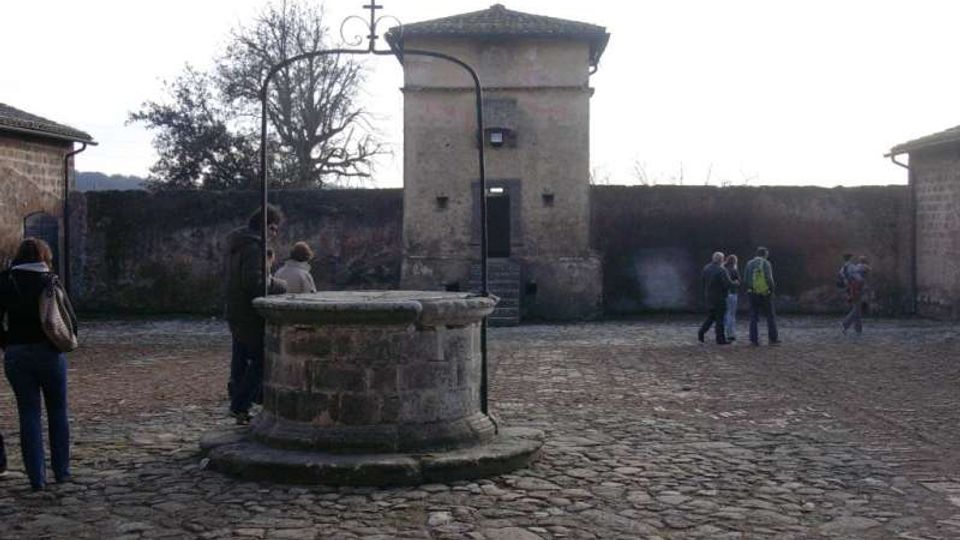 Cortile lastricato in pietra con un grande pozzo circolare in primo piano, dotato di bracci metallici per la carrucola. Sullo sfondo si vede un edificio in muratura con porta centrale e tetto a padiglione, affiancato da mura perimetrali e altri fabbricati.
