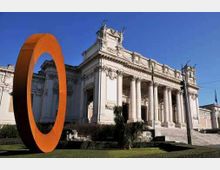 Facciata di un edificio monumentale in stile neoclassico con grande portico colonnato e scalinata in pietra, sotto un cielo sereno. In primo piano, su un prato, è visibile una grande scultura anulare arancione.