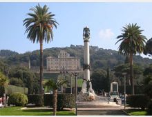Piazza con giardini curati e alte palme, con una colonna monumentale e fontana al centro. Sullo sfondo, su una collina boscosa, si vede un grande edificio storico in stile palazzo.