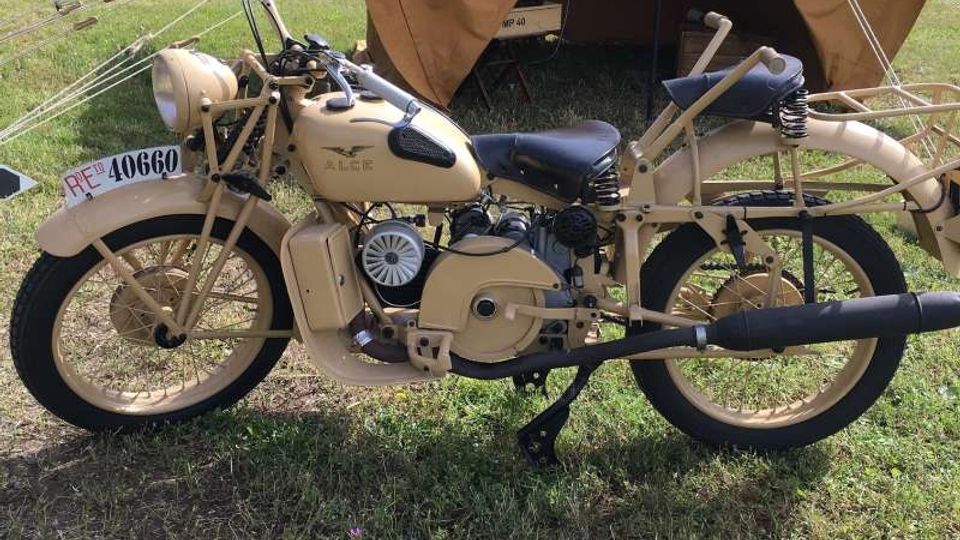 Motocicletta d’epoca color beige parcheggiata su un prato, con cavalletto e ruote a raggi in evidenza. Sullo sfondo si vede una tenda da campo in tela marrone montata in un’area verde, con alberi e altre strutture lontane.