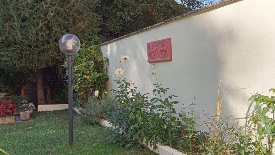 Giardino residenziale con prato verde, aiuole di fiori rossi e bianchi e una lampada da esterno a globo. Sul lato destro c’è un lungo muro chiaro con una targhetta, bordato da piante e fiori, con alberi alti sullo sfondo sotto un cielo sereno.