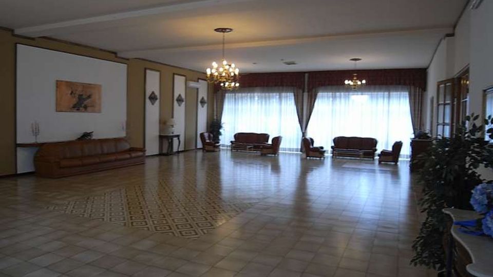 Ampia sala interna con pavimento in piastrelle e lampadari a soffitto, arredata con divani e poltrone disposti lungo le pareti. Grandi finestre con tende e drappeggi occupano il fondo della stanza, con elementi decorativi e piante ai lati.