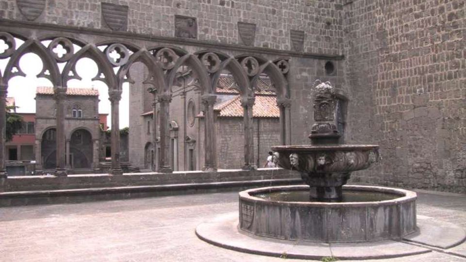 Cortile in pietra con una grande fontana circolare al centro, circondato da alte mura in blocchi di tufo. Sul lato sinistro si vede una loggia con archi gotici e colonnine che incorniciano edifici storici sullo sfondo.