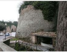 Muro di fortificazione in pietra con struttura semicilindrica e vegetazione sulla sommità, lungo una strada con lampione e auto parcheggiate. In primo piano si vede un piccolo edificio con tetto in coppi e una recinzione in legno su un percorso pedonale.