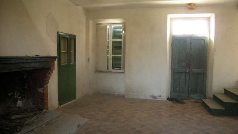 Interno di una stanza spoglia con pareti intonacate chiare e pavimento in mattoni, con grande camino in muratura su un lato. Sono visibili una finestra e due porte in legno verde, una delle quali rialzata su alcuni gradini.
