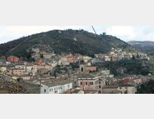 Panorama di un borgo collinare con case addossate su un pendio, tetti in coppi e un campanile in primo piano. Sullo sfondo si vede una grande collina boscosa con strade serpeggianti e alcune strutture sulla cresta.
