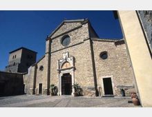 Facciata in pietra di una chiesa storica con frontone triangolare, rosone circolare e portale decorato con architrave scolpito. Davanti si apre una piazza lastricata; sullo sfondo a sinistra si vede una torre in pietra sotto un cielo sereno.