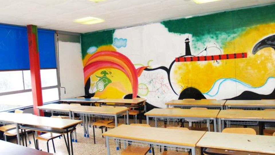Aula scolastica con file di banchi e sedie in legno su pavimento in graniglia, illuminata da plafoniere a soffitto e con grandi finestre schermate da tende blu. La parete di fondo è decorata con un ampio murale colorato che raffigura forme astratte, un arcobaleno e un faro su uno sfondo giallo e verde.