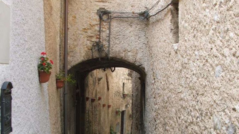 Vicolo stretto in pietra tra edifici in muratura, con pavimentazione irregolare e un piccolo arco che collega le pareti sopra il passaggio. Sulle facciate si vedono cavi e tubazioni a vista, una finestra piccola e alcuni vasi appesi.