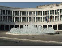 Ampia piazza con una grande fontana circolare a getti d’acqua al centro, davanti a un edificio moderno curvilineo in pietra chiara con portici e molte finestre. Sulla facciata sono visibili alcune bandiere vicino all’ingresso.