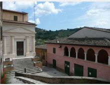 Piazza in un borgo collinare con una chiesa chiara a facciata classica e scalinata in pietra. Di fronte si vede un edificio rosa con portico ad archi e tetto in coppi, sullo sfondo colline verdi e cielo parzialmente nuvoloso.