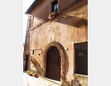 Facciata di un edificio antico in intonaco color ocra, con grande portale ad arco in pietra e portone in legno. Si vedono finestre con grate, cavi esterni lungo il muro e una strada lastricata in pietra con vasi di fiori.
