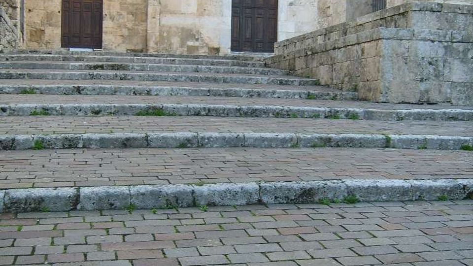 Scalinata in pietra che conduce a una facciata storica in blocchi di pietra chiara, con due portali ad arco e porte in legno scuro. A destra si vede un grande arco d’ingresso con parapetto in pietra e una finestra stretta sopra uno dei portali.
