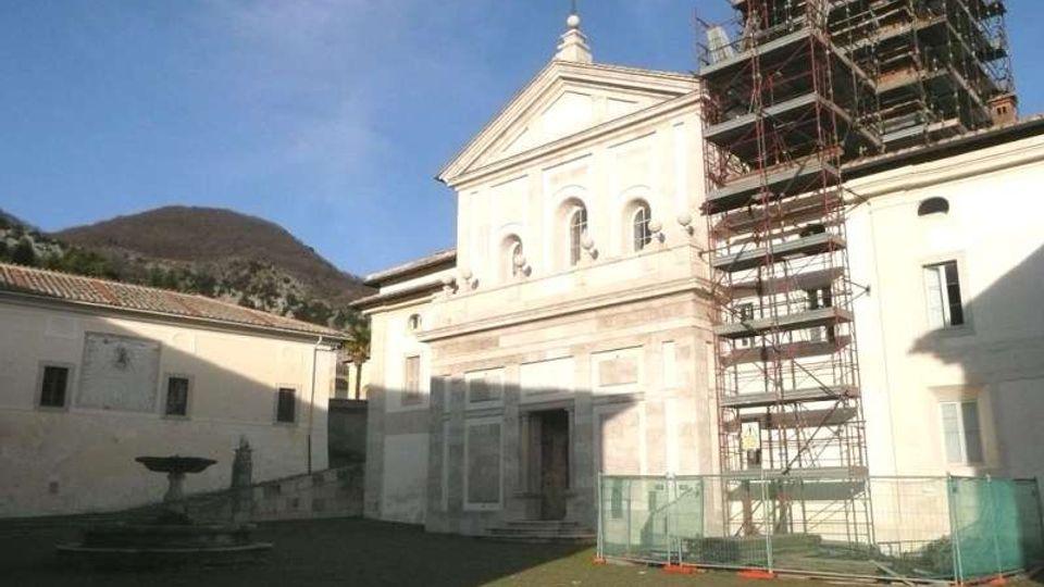 Facciata di una chiesa in una piazza con pavimentazione in pietra, con frontone triangolare e tre finestre ad arco; sul lato destro è presente un’alta impalcatura da cantiere con recinzione. Sullo sfondo si vedono edifici chiari e una collina sotto un cielo sereno.