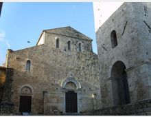Facciata in pietra di una chiesa medievale con portali ad arco e piccole finestre, affacciata su una piazza. A destra si vede una torre campanaria in pietra con grande arco e apertura superiore, sullo sfondo di cielo sereno.