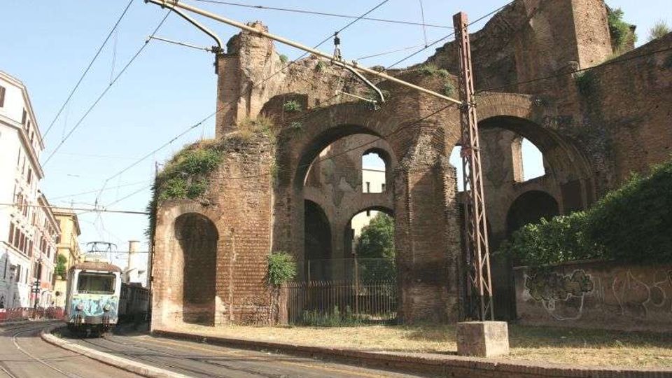 Ruderi di un antico acquedotto in mattoni con grandi arcate sovrapposte, accanto a una strada urbana. In primo piano si vedono i binari e la linea elettrica del tram, con edifici ai lati e vegetazione che cresce sulle mura.