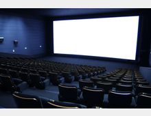 Sala cinematografica con file di poltrone scure disposte in gradinata e rivolte verso un grande schermo luminoso. Pareti e soffitto scuri con illuminazione discreta laterale e altoparlanti montati sulle pareti.