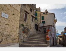 Scalinata in pietra in un borgo storico, affiancata da case in muratura con persiane verdi e balconi. Lungo i gradini ci sono fioriere con fiori rossi, con una stretta via che scende sulla destra e un’insegna “BAR” visibile sullo sfondo.