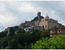 Veduta di un borgo collinare con case in pietra e intonaco addossate al pendio, circondate da vegetazione. In cima spiccano una torre cilindrica medievale e una chiesa con campanile sotto un cielo nuvoloso.