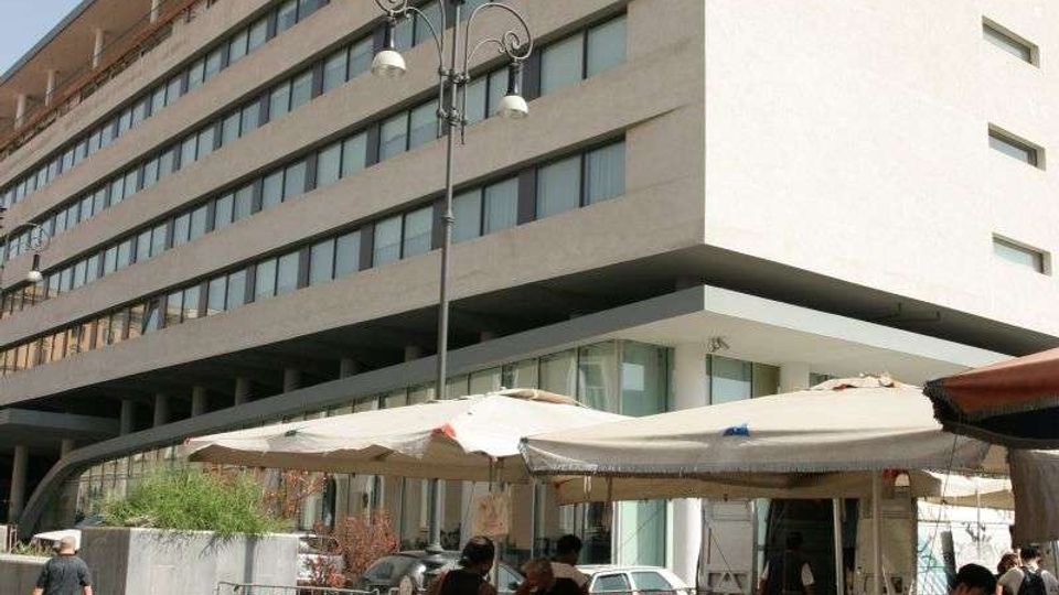 Edificio moderno a più piani con facciata chiara e lunghe finestre orizzontali, visto da un angolo su una strada urbana. In primo piano c’è un mercato all’aperto con bancarelle sotto grandi tende bianche, cassette di frutta e verdura e un lampione centrale.
