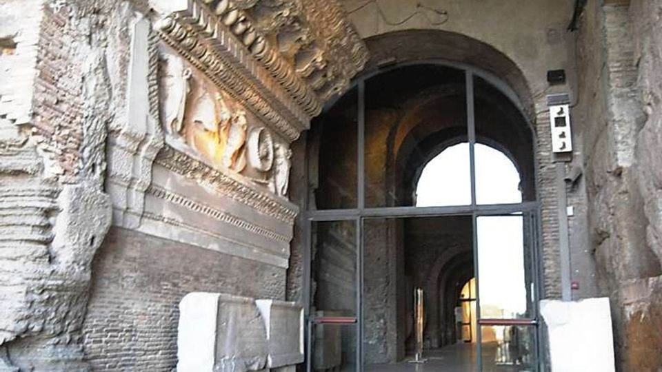 Ingresso di un antico edificio in muratura con grande arco e volte in pietra, pavimento in lastre e portale moderno in vetro e metallo. Sulla parete sinistra si vede un fregio decorativo sporgente e blocchi di pietra esposti lungo il corridoio.