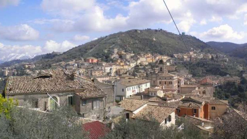 Veduta panoramica di un borgo collinare con case in pietra e tetti in coppi, disposto su pendii terrazzati. Sullo sfondo si vedono colline boscose e un cielo parzialmente nuvoloso.