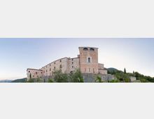 Grande complesso in pietra tipo castello o monastero su un basamento murario, con torre cilindrica e finestre ad arco, su una collina con vegetazione e montagne sullo sfondo sotto un cielo sereno.