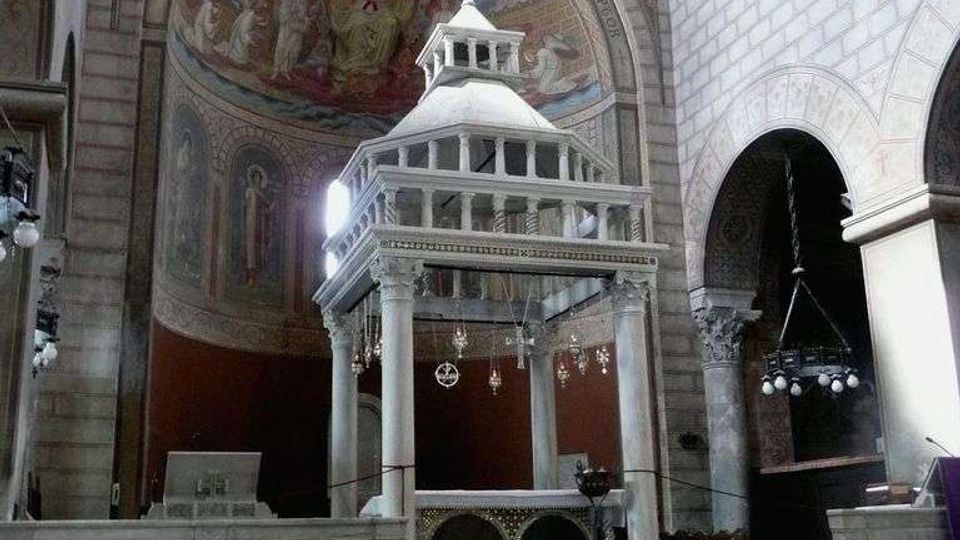 Interno di una chiesa in pietra con grande abside affrescata e arcate laterali; al centro un ciborio marmoreo a colonne sopra l’altare, con pavimento a motivi geometrici e copertura a capriate lignee.