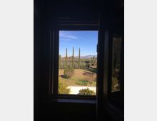 Vista attraverso una finestra rettangolare in un interno scuro, che incornicia un paesaggio di campagna con prato, alberi e alti cipressi. Sullo sfondo si vedono colline o montagne sotto un cielo sereno.