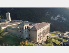 Complesso monastico in pietra su un pendio, con edifici a tetti in coppi e un alto campanile quadrato. Davanti si vedono un cortile e giardini terrazzati, con una collina boscosa sullo sfondo.