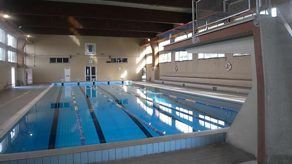 Piscina coperta con vasca lunga a corsie delimitate da galleggianti, acqua azzurra e bordo piastrellato. L’ambiente è un grande locale con travi a vista sul soffitto, ampie finestre laterali e una passerella sopraelevata con ringhiera metallica su un lato.