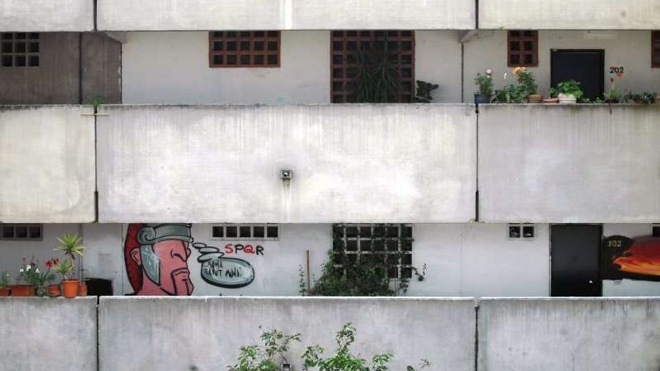 Facciata di un edificio residenziale in cemento con lunghi balconi sovrapposti, ringhiere piene e piccole finestre a griglia. Sui balconi si vedono vasi con piante e un murale/graffito colorato su una parete.