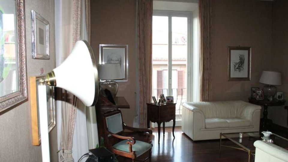 Soggiorno arredato con divani chiari, poltrona e tavolino su pavimento in legno scuro, con quadri alle pareti e lampade da tavolo. Al centro una portafinestra con tende apre su un balcone con vista su edifici esterni.