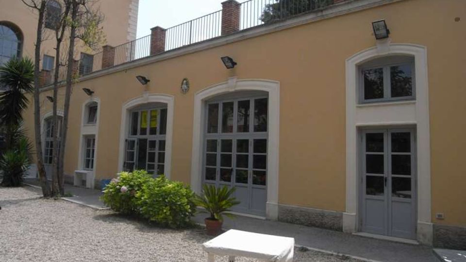 Cortile con ghiaia davanti a un edificio basso color ocra, con grandi porte e finestre ad arco incorniciate di bianco e infissi grigi. Sulla facciata ci sono lampade a parete, una ringhiera sul tetto e alcune piante e arbusti in vaso vicino all’ingresso.