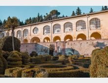 Edificio storico intonacato con porticato ad archi e grandi finestre ad arco, affacciato su un muro in pietra. In primo piano un giardino formale con siepi potate in forme geometriche e un’alta palma, con alberi sullo sfondo.
